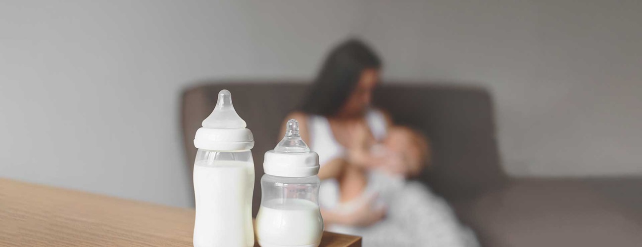 A collection of milk bottles sit on a table, as a mother breastfeeds her baby in the background.