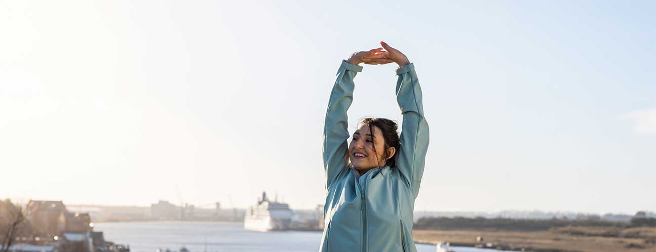 Woman in winter coat stretches before exercising. In the background sits a peaceful river with several ships. 