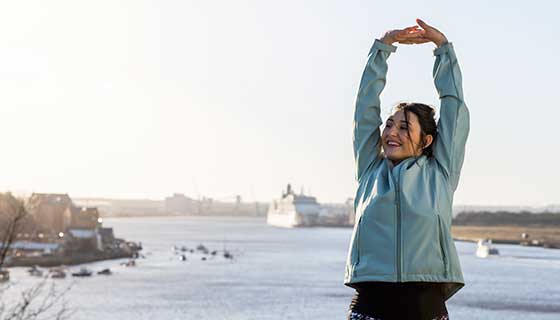 Woman in winter coat stretches before exercising. In the background sits a peaceful river with several ships. 