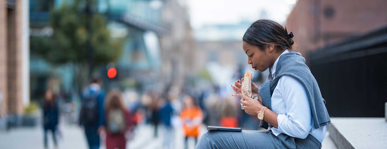 A woman sitting on the steps of a building and eating a wrap while looking at her tablet