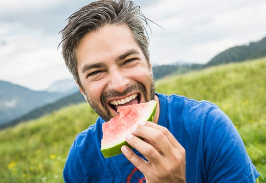 A middle aged man eating a watermelon slice in the middle of a field. 
