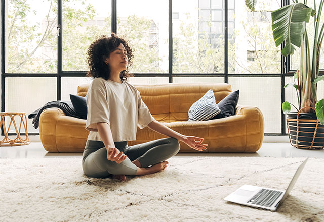 A woman meditating in her living room, in front of her laptop.