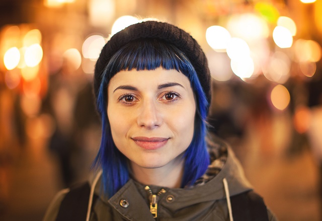 young woman with blue hair and beanie at foreground, lights out of focus in background.