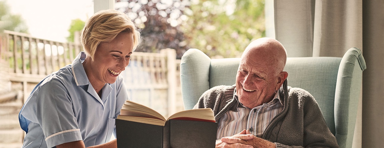 A caregiver reads a book with an elderly patient in their home.