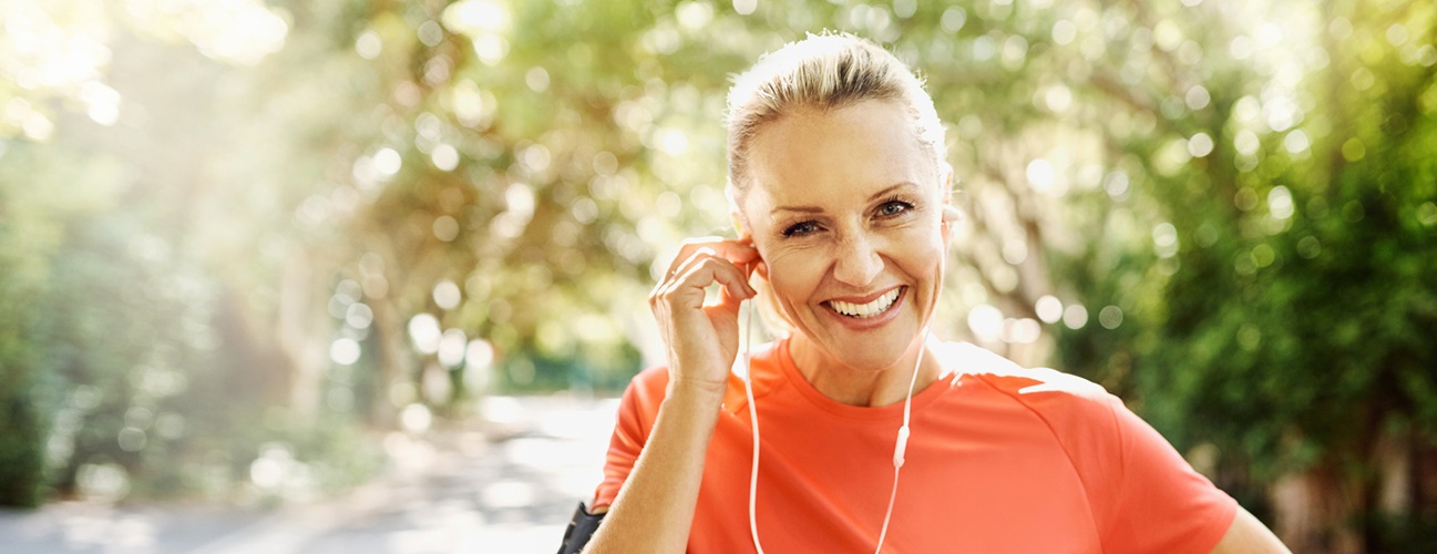 A woman stands outside, smiling, as she adjusts her headphones. She is enjoying an active lifestyle following her breast reconstruction.