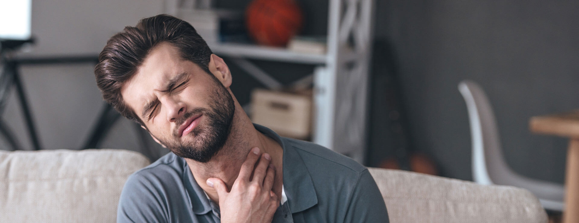Frustrated handsome young man touching his neck and holding a glass of water while sitting on the couch at home