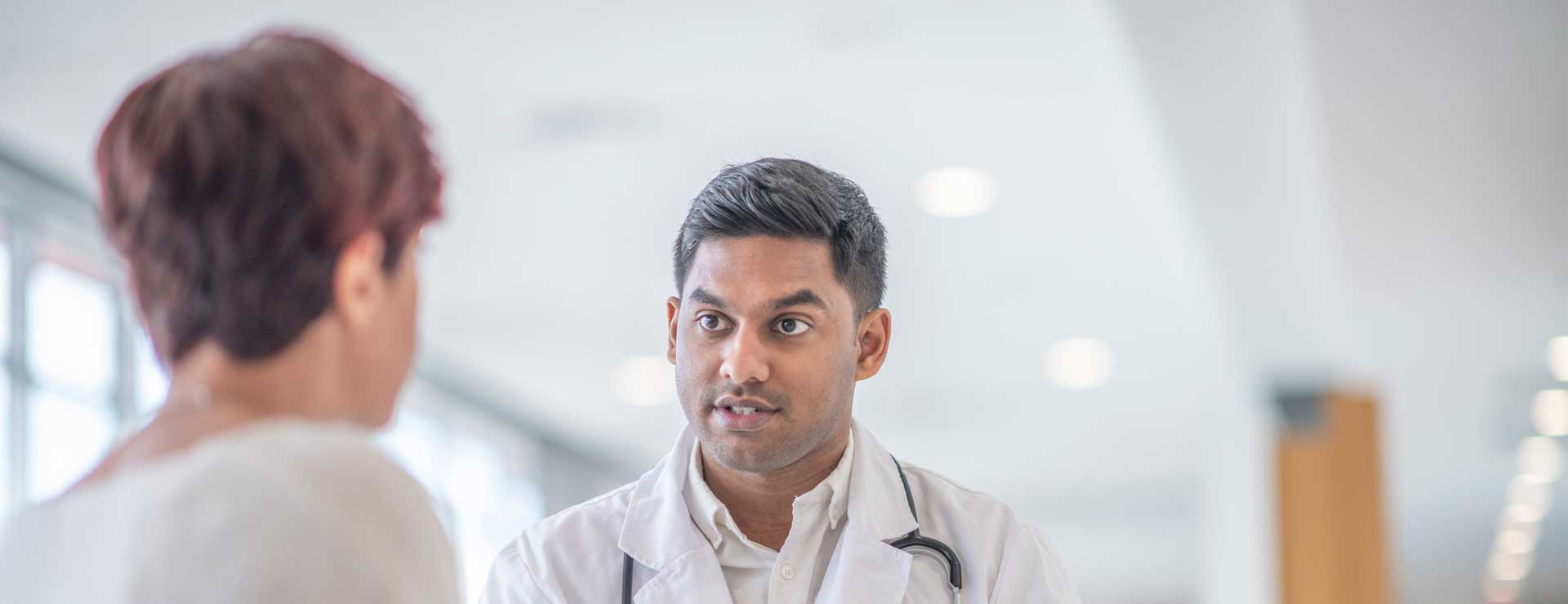 A female adult is at a check up appointment for a diabetic medical condition. She is sitting across the table from her Indian doctor who is holding a glucometer pen that is used to test her blood sugar levels.