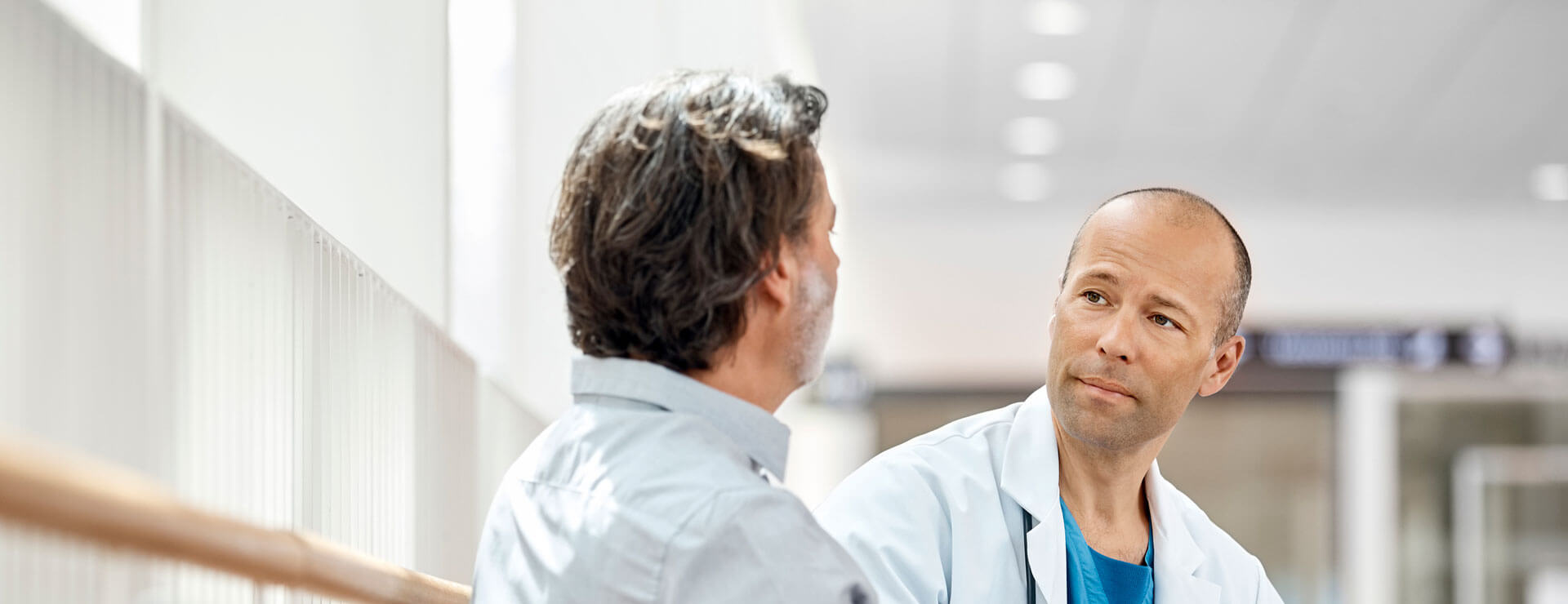 Male doctor counseling mature patient in waiting room. Medical professional is listening to ill man while holding digital tablet in hospital. They are sitting on seat.