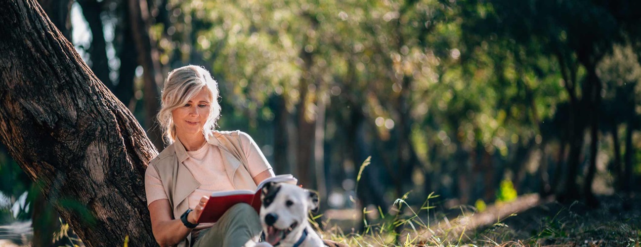 Older woman relaxes in forest with book and dog.