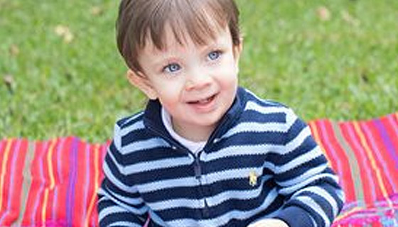 Young boy sitting on a blanket in the grass, smiling. 