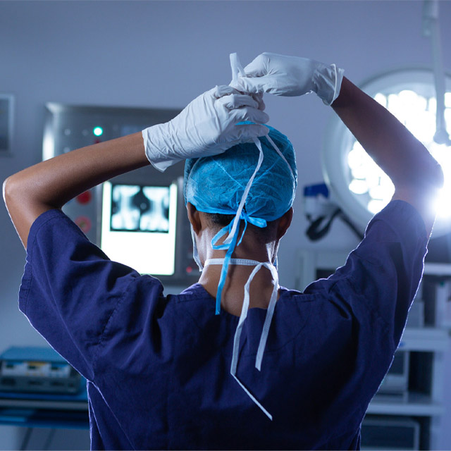  A surgeon, seen from the back, wearing navy blue scrubs and a surgical cap, ties the strings of their surgical mask in an operating room.