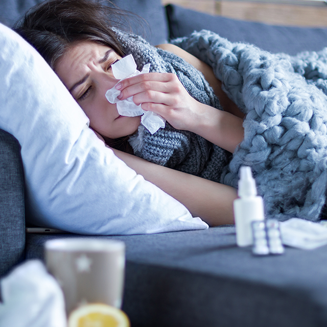 An exhausted, sick young woman lying on a couch wrapped in a gray blanket blowing her nose.