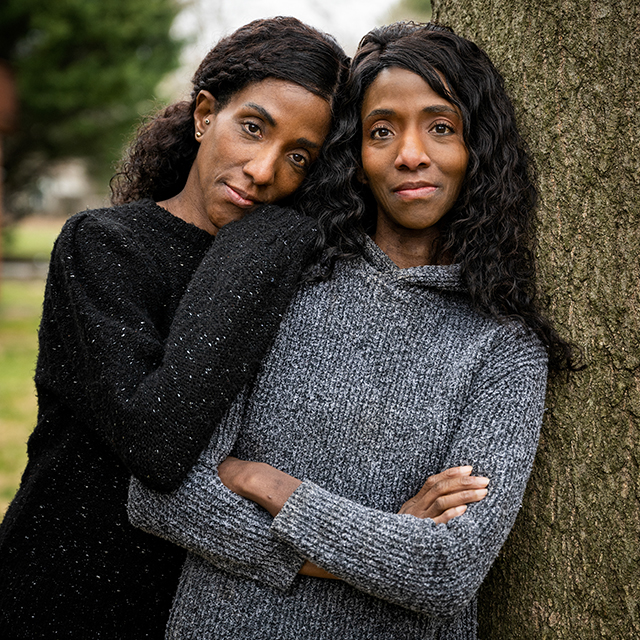 A pair of twins lean against a tree. One wears a black sweater while the other wears a grey sweater. 