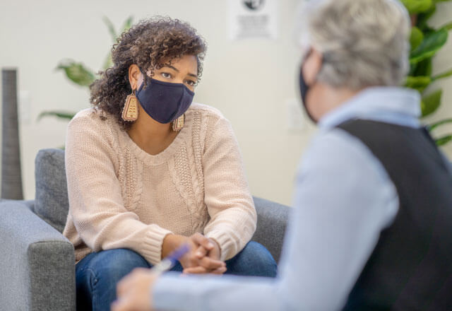 A masked medical professional sits listening to their patient.