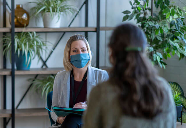 A masked medical professional sits with a clipboard, listening to their patient.