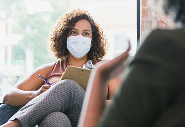 A masked medical professional sits with a clipboard, listening to their patient.