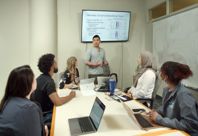 students sitting around a conference table looking at a student standing at the head of the table presenting from a screen behind them