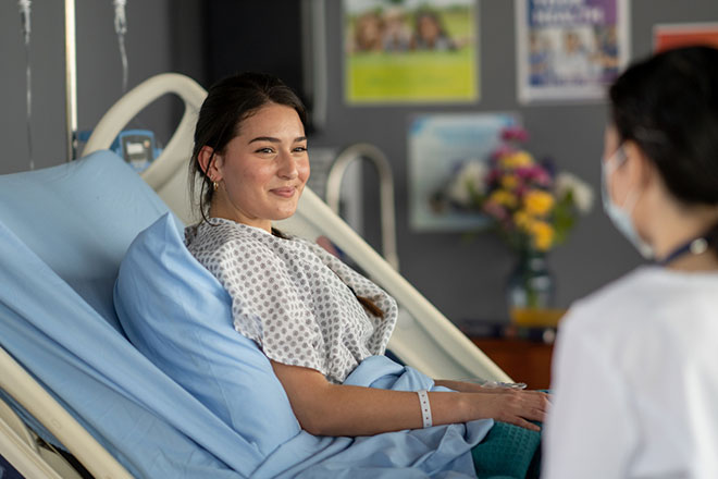 A teenager in a hospital bed talks with her nurse