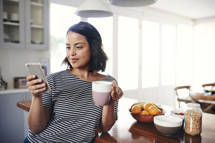 woman looking at her phone in kitchen while drinking out of a mug