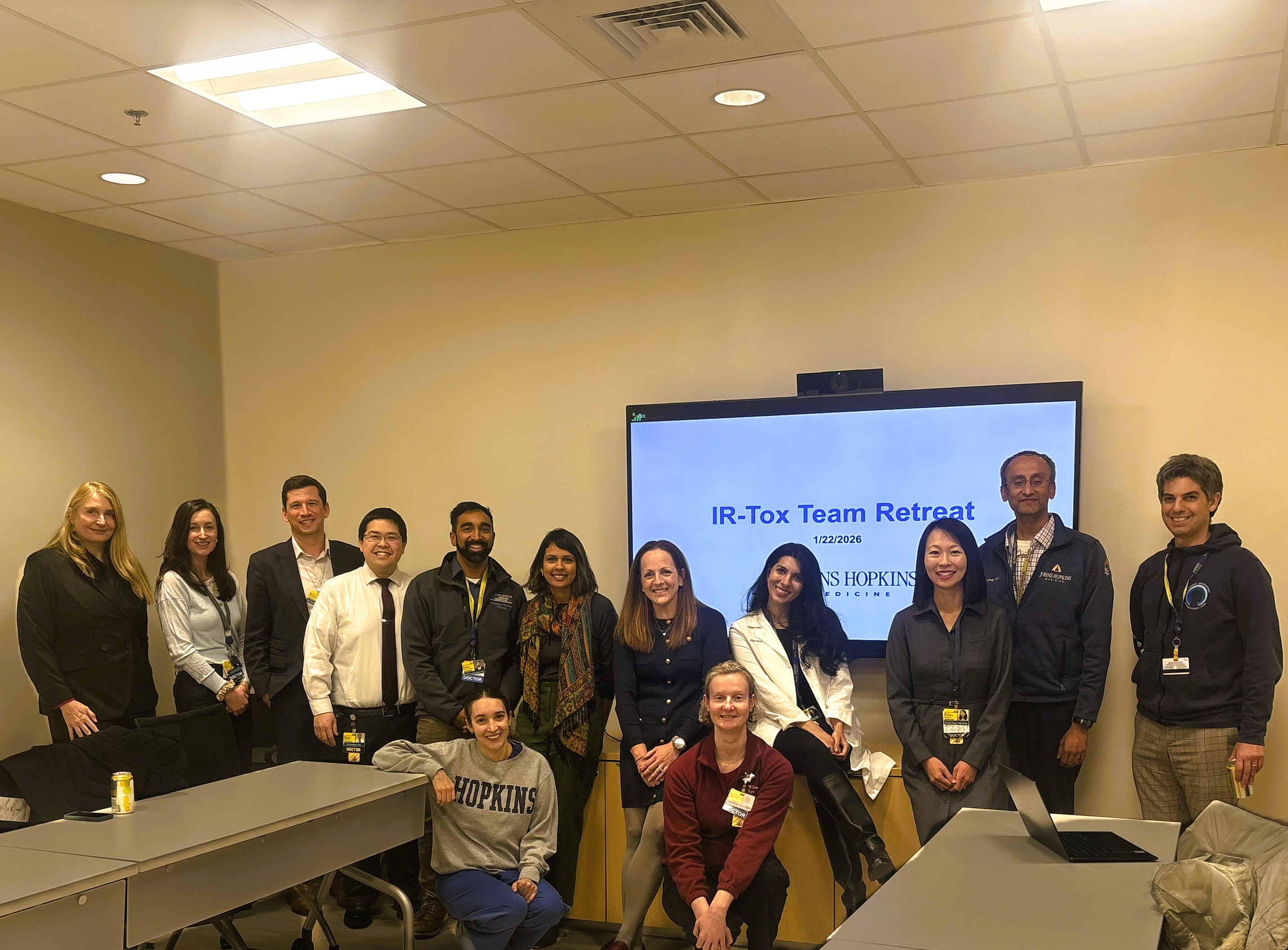 Group photo of the IR‑Tox team gathered in a conference room during a retreat, standing and seated in front of a screen displaying “IR‑Tox Team Retreat” with the Johns Hopkins Medicine logo; laptops and tables are visible in the meeting space.