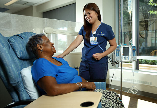 Female middle aged patient sitting in cushioned leather chair, smiling at a nurse who is smiling back at her, while standing next to her with her hand on the patient's shoulder.