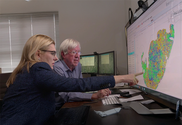 Two people examine a large map on a computer screen, pointing to colorful data points. The room is modern, with multiple monitors, conveying focus and collaboration.