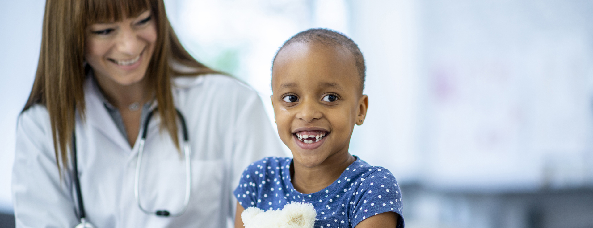 Pediatric patient holding a white teddy bear smiling with doctor smiling down at her, both seated next to each other. 