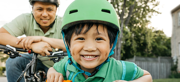 A boy and his father riding bikes together. The boy beams a big smile right front and center, prominently wearing his green safety helmet.