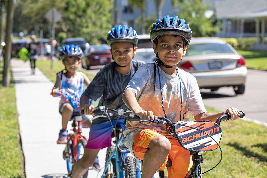 A group of children riding their bikes outside on the sidewalk while wearing bicycle helmets