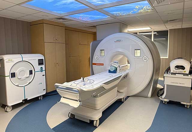 MRI machine room with modern equipment, featuring a large scanner and a sky-themed ceiling panel. The atmosphere is clinical yet calming.