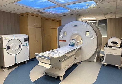 MRI machine room with modern equipment, featuring a large scanner and a sky-themed ceiling panel. The atmosphere is clinical yet calming.