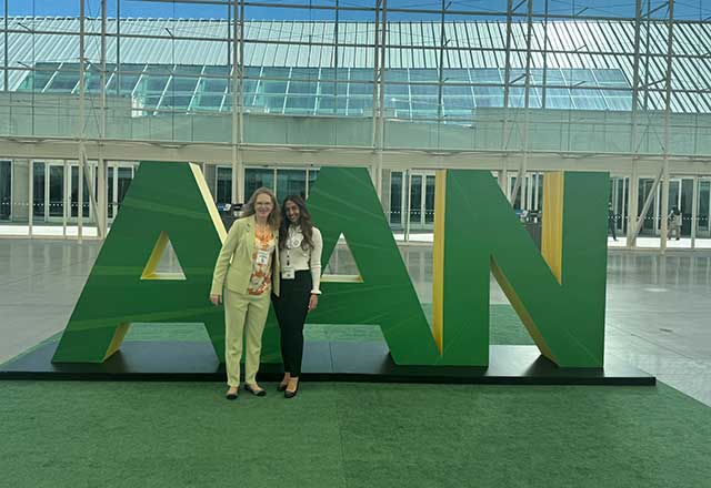 Dr. Muehlschlegel and a female trainee stand in front of large green and yellow "AAN" letters inside a spacious, modern building with glass walls.