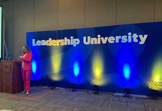 Dr. Muehlschlegel in a red suit speaks at a podium beside a large "Leadership University" sign with blue and yellow lights in the background.