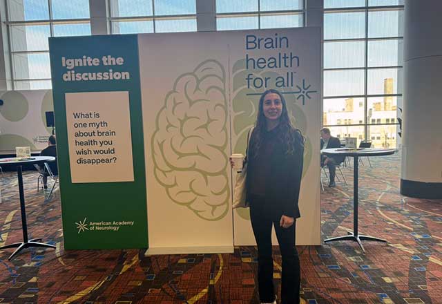 A woman stands in front of a display about brain health at a conference, holding a drink and smiling at the camera. The display features a large brain illustration and informational text.