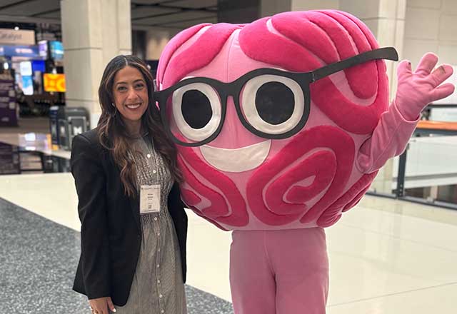 A woman stands next to a person in a large pink brain mascot costume with glasses, smiling and waving inside a convention center.