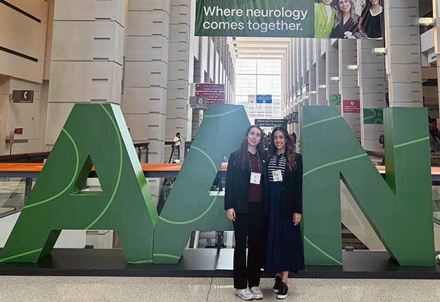 Two women stand smiling in front of a large sign displaying the word "AAN."