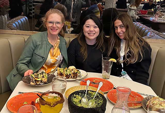 Three women sit at a restaurant booth, smiling. The table has plates of food, colorful dishes, and drinks, creating a lively and cheerful atmosphere.