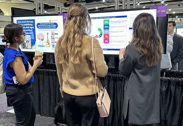 Three people stand closely viewing a presentation poster displaying graphs and data on live music therapy. The mood is focused and engaged.