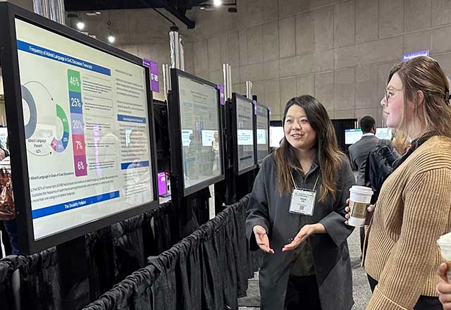 Two women discuss a scientific poster at a conference. One gestures while explaining data on a screen. Both appear engaged and focused.