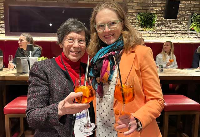Two women smile at the camera while holding cocktail glasses filled with orange drink and straws in a restaurant setting.