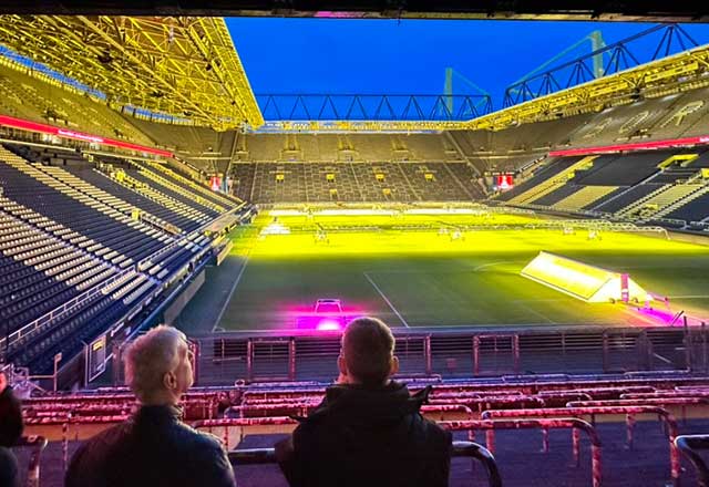 View of a brightly lit football stadium at twilight, showing two seated figures and a vibrant green field illuminated in yellow and pink.