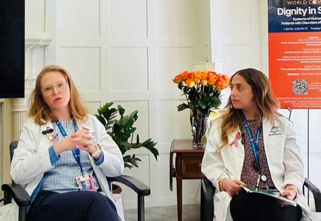 Two women in lab coats engaged in conversation while seated in chairs in a conference setting.
