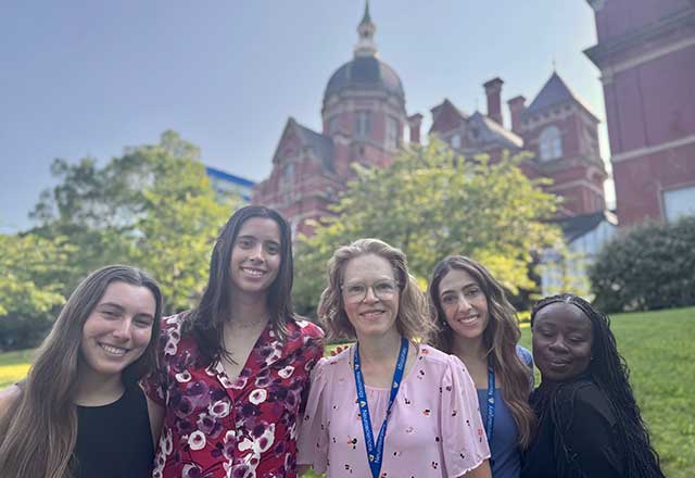 A group of five smiling women from the Muehlschlegel Lab stand outside on a sunny day with the Johns Hopkins dome building and trees in the background, conveying a sense of joy and camaraderie.