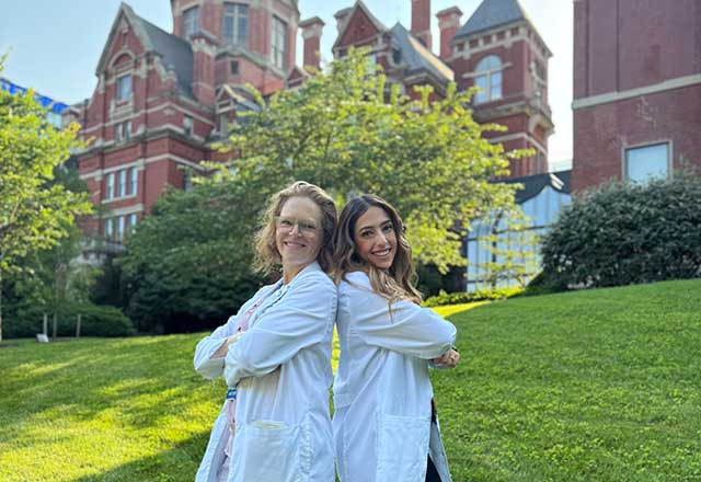 Two women in white lab coats stand back-to-back, smiling confidently, in front of the historic Johns Hopkins dome building with lush greenery and a sunny sky.