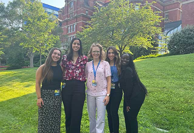 A group of five women stands smiling on a sunny lawn in front of a red brick building and trees, conveying a cheerful and collegial atmosphere.