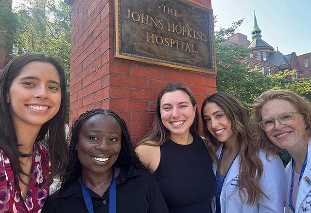 Five women smiling in front of The Johns Hopkins Hospital sign, set against a backdrop of brick buildings and greenery, conveying a positive and welcoming tone.