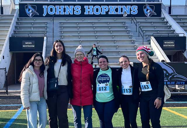 A group of smiling women in warm clothing, some wearing bibs with racing numbers, pose on a stadium field, with Johns Hopkins sign and bleachers in the background.