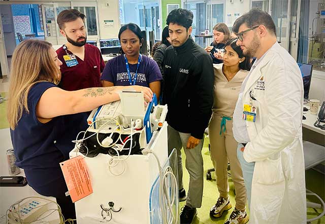 A group of NCC fellows and Dr. Shah attentively watch a demonstration on medical equipment in a hospital setting, conveying focus and learning.