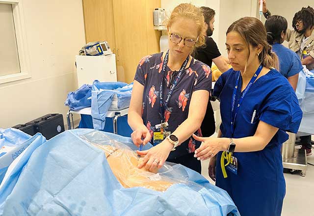 Two medical professionals in scrubs focus intently on a covered medical dummy in a hospital room, demonstrating a procedure. The room is busy with other staff.
