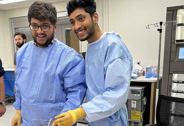 Clinical fellows in blue gowns and yellow gloves smile while practicing a procedure on a medical dummy in a clinical setting.